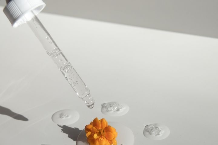 From above of drops of transparent moisturizing cosmetic product dripped by pipette and small fresh flower head placed on white table